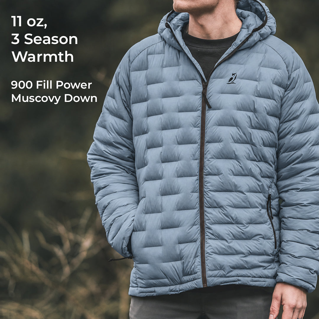 A man wearing a light blue hooded puffer jacket with a full-zip front and horizontal quilted baffles, shown outdoors against a blurred natural background. White text on the left reads '11 oz, 3 Season Warmth' and '900 Fill Power Muscovy Down'.