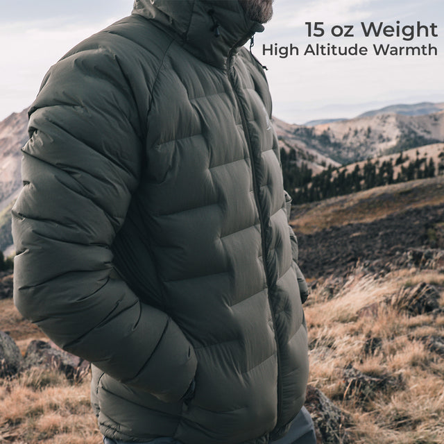 A man wearing a dark puffer jacket stands outdoors in a mountain landscape. Text in the upper right reads '15 oz Weight High Altitude Warmth'.