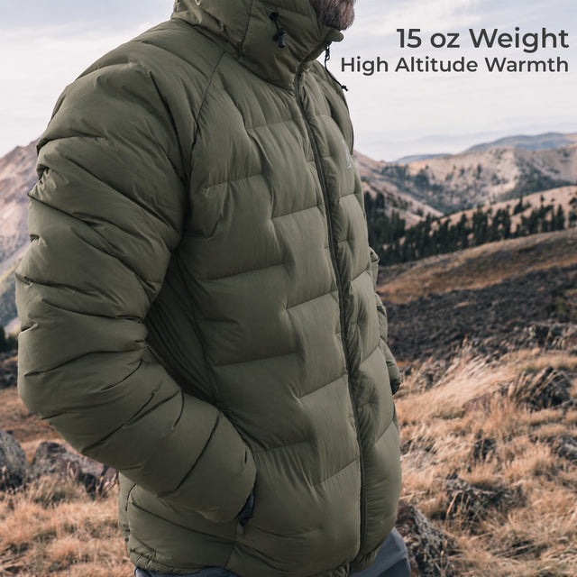 A man wearing a dark olive puffer jacket stands outdoors in a mountain landscape. Text in the upper right reads '15 oz Weight High Altitude Warmth'.