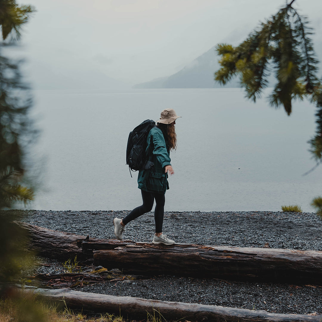 A hiker wearing a sun hat balancing on a fallen log along a misty lake shoreline while wearing the Outdoor Vitals KotaUL Ultralight Travel & Adventure Backpack.