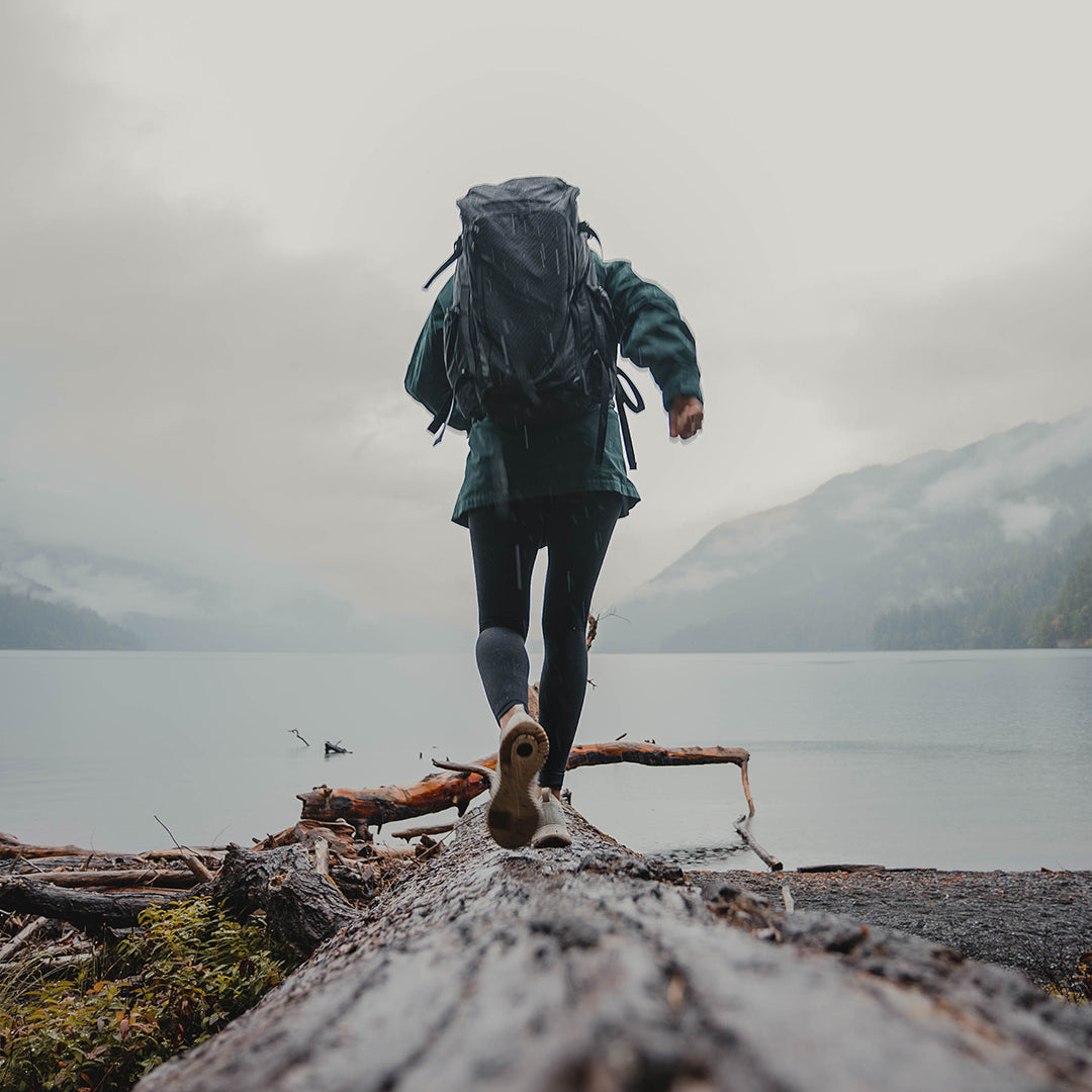 Rear view of a person wearing the Outdoor Vitals KotaUL Ultralight Travel & Adventure Backpack while balancing on a large log along a foggy lake shore with mountains in the background.