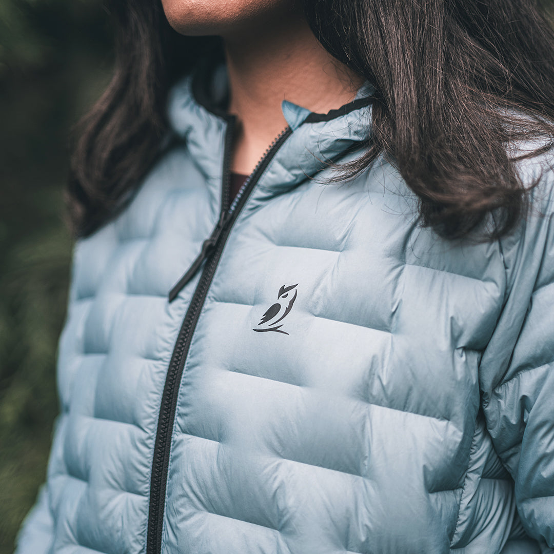 A close-up of a person wearing a light blue puffer jacket outdoors. The jacket has a black zipper and a small black bird logo on the left chest.