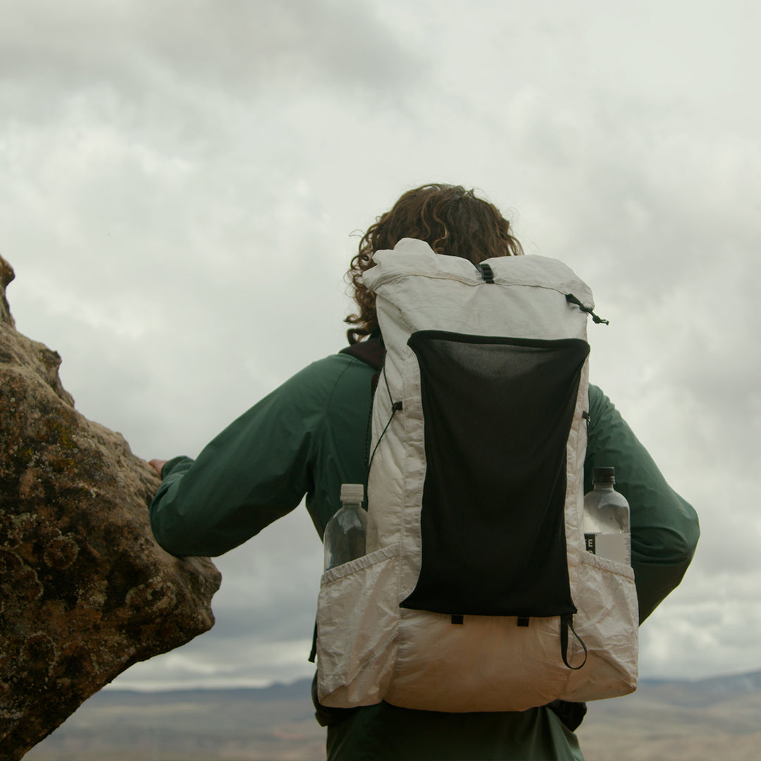Back view of a hiker resting against a rock while wearing the Outdoor Vitals CS40 Ultra Backpack, highlighting its large front stretch mesh pocket, roll-top closure, and water bottle side pockets.