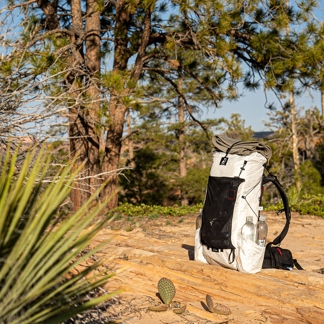 Fully loaded Outdoor Vitals CS40 Ultra Backpack sitting upright on sandstone terrain among desert pines, featuring dual water bottles, front mesh pocket, and top-strapped foam pad.