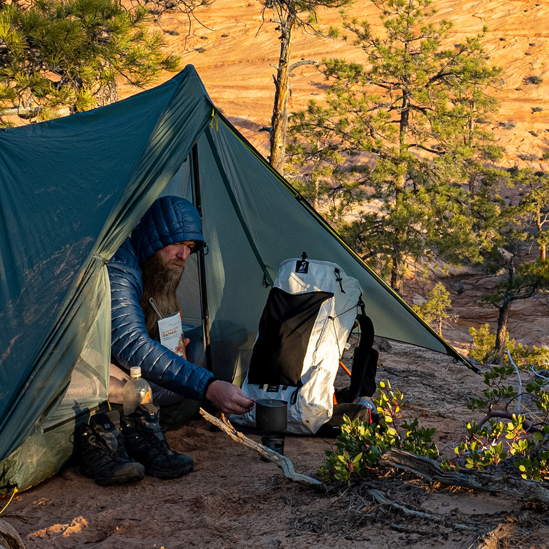 A bearded camper resting inside a trekking-pole tent while preparing food next to the Outdoor Vitals CS40 Ultra Backpack at a backcountry campsite in a canyon landscape.