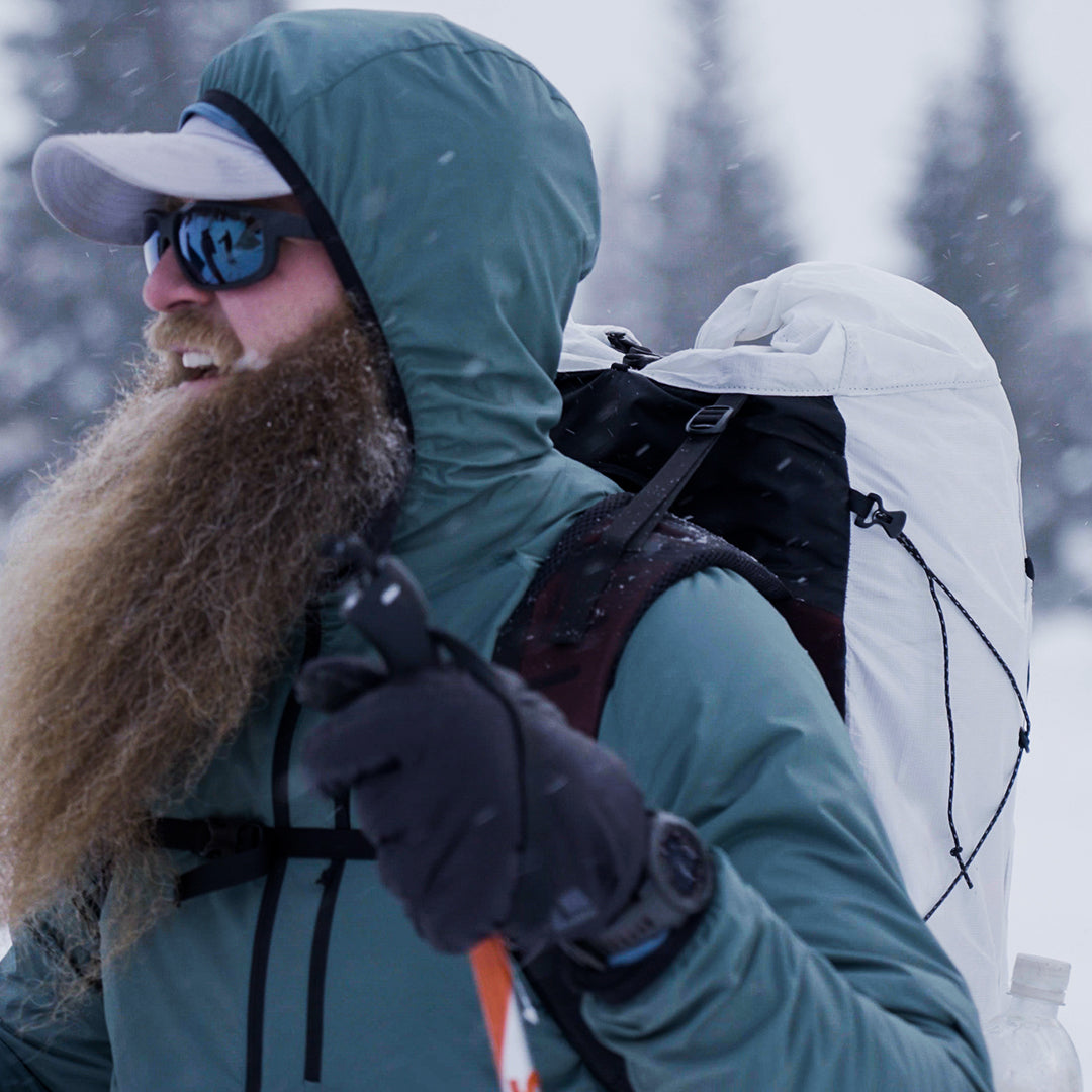 Side profile of a bearded winter hiker smiling while wearing a hooded jacket, cap, and the Outdoor Vitals CS40 Ultra Backpack during a snowstorm on a mountain trail.