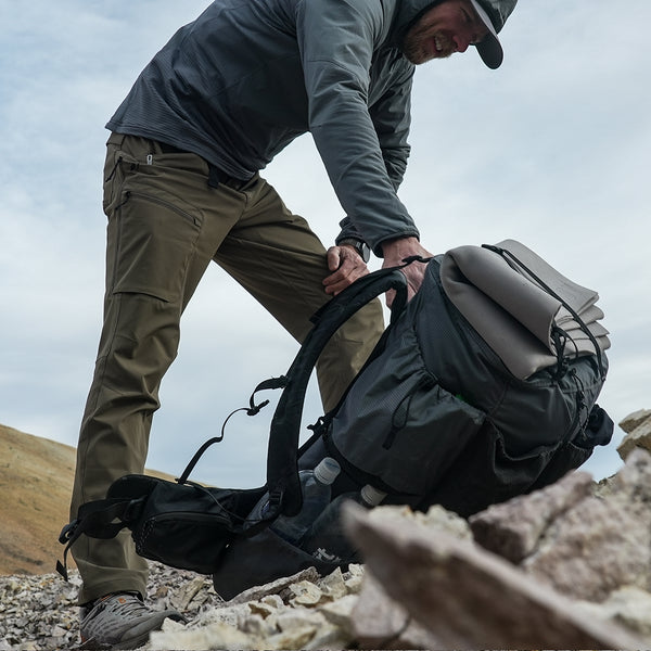 side view of man picking up backpack on mountain ridge - hiking pants and hoodie are visible