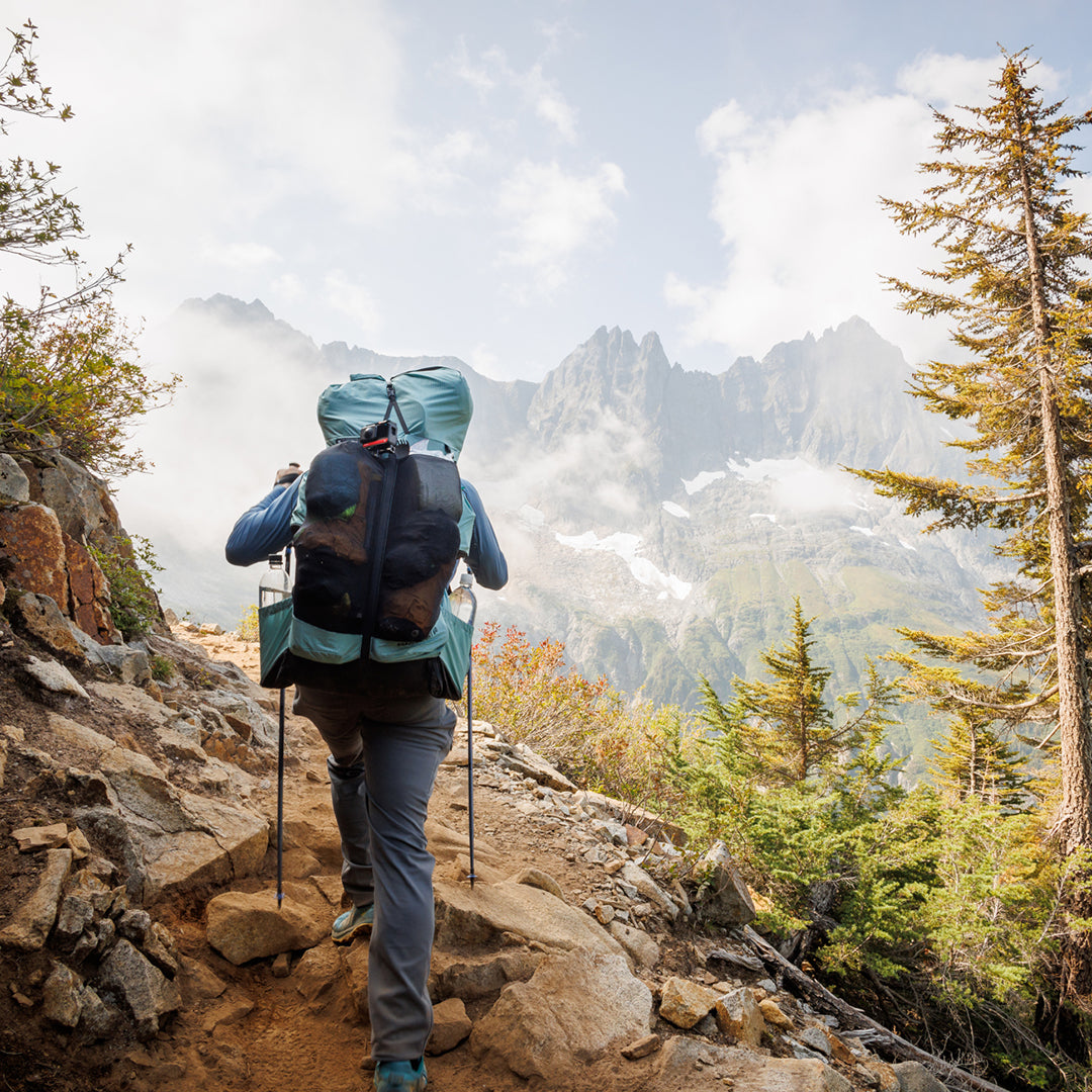Rear view of a hiker ascending a rugged mountain trail carrying the Outdoor Vitals Shadowlight Carbon 60 Backpack, featuring large rear mesh storage and side water bottle pockets.