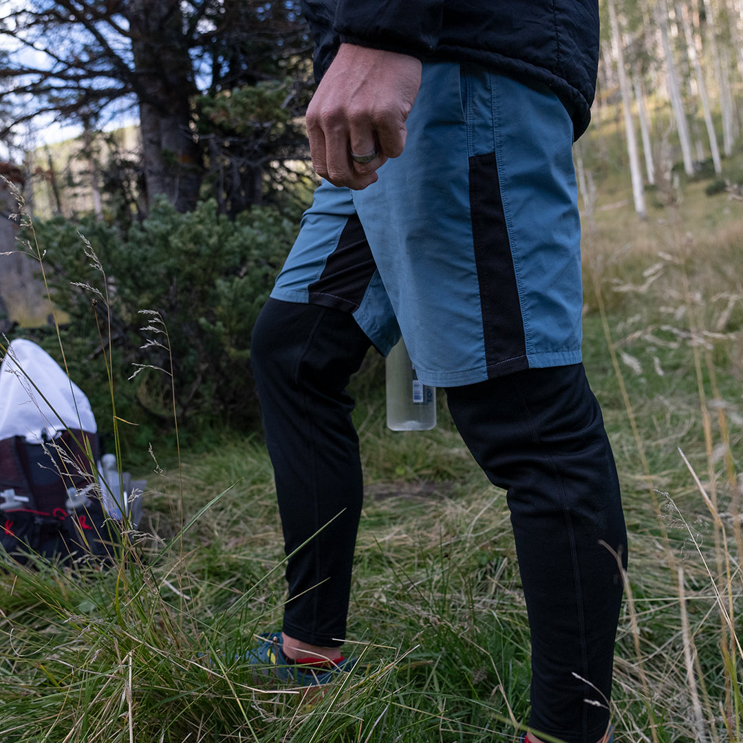 Lower-body view of a male hiker wearing Outdoor Vitals Highline Thermal Leggings layered under athletic shorts while standing in a grassy outdoor environment.