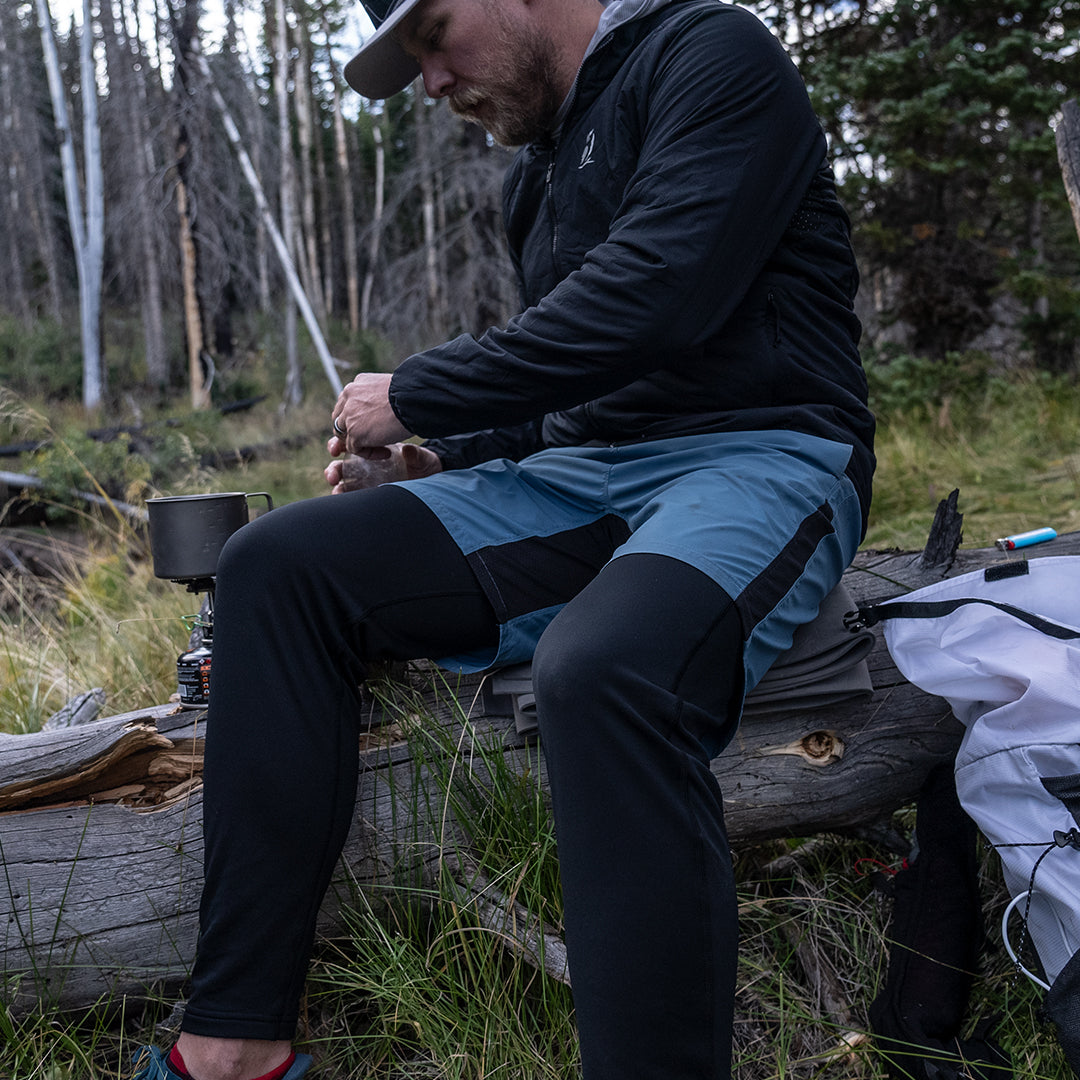 A male hiker sitting on a fallen log while preparing food at a campsite, wearing Outdoor Vitals Highline Thermal Leggings layered under athletic shorts.