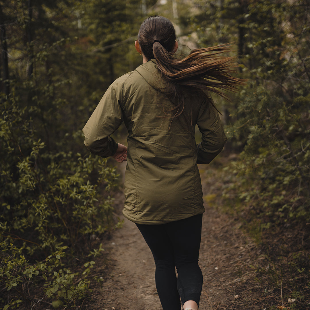 Back view of a woman trail running in a Ventus Active Hoodie through a wooded forest path.