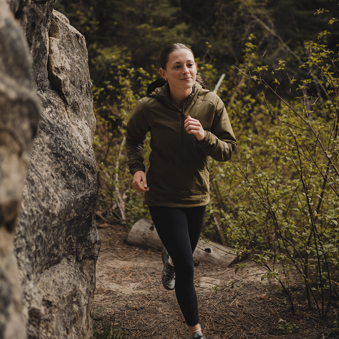 Woman running on a wooded trail next to a rock wall, wearing an Ventus Active Hoodie and leggings.
