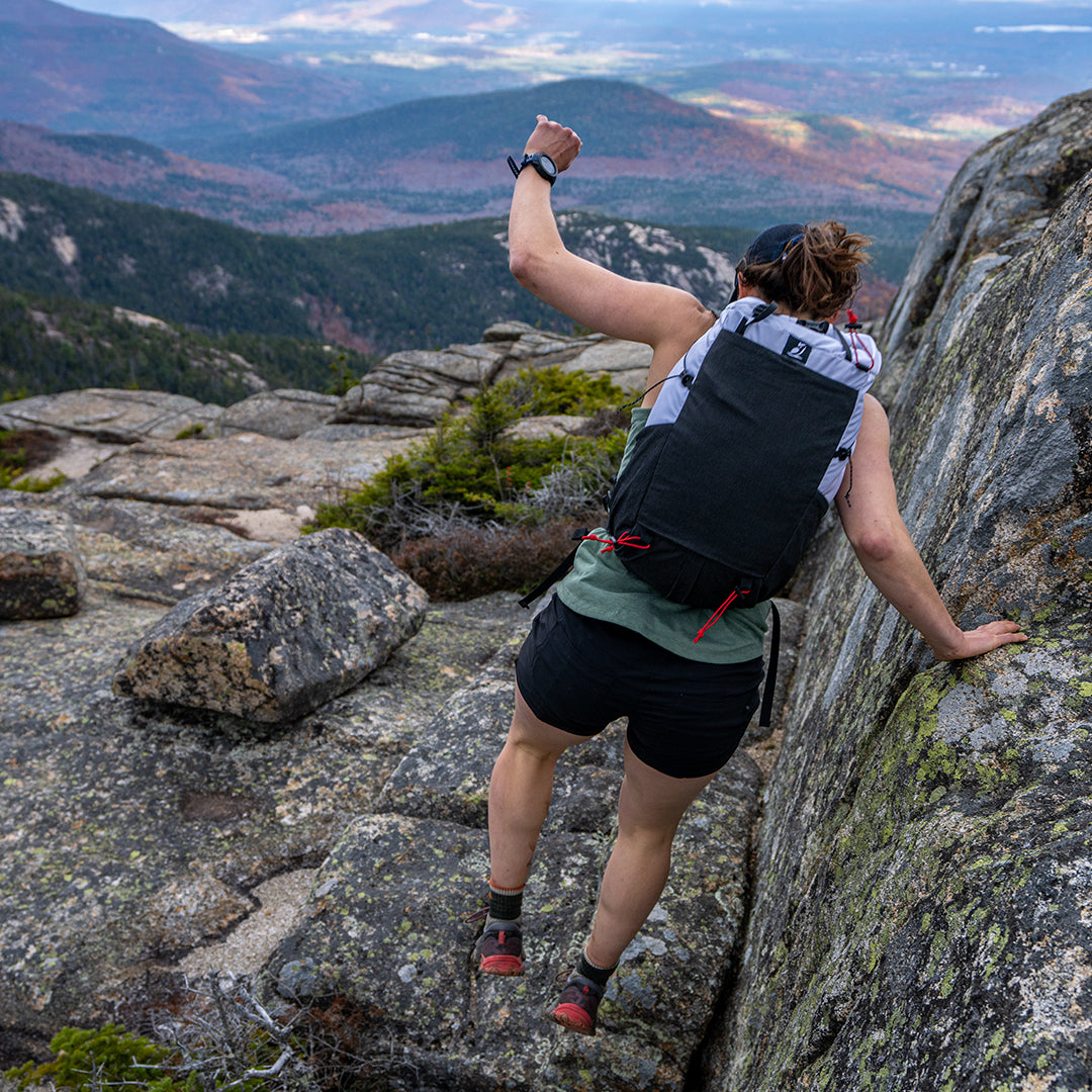 Rear view of a hiker scrambling over rocky mountain terrain wearing the Outdoor Vitals Skyline 30 Fastpack, showing the secure fit during technical movement and scenic valley views below.