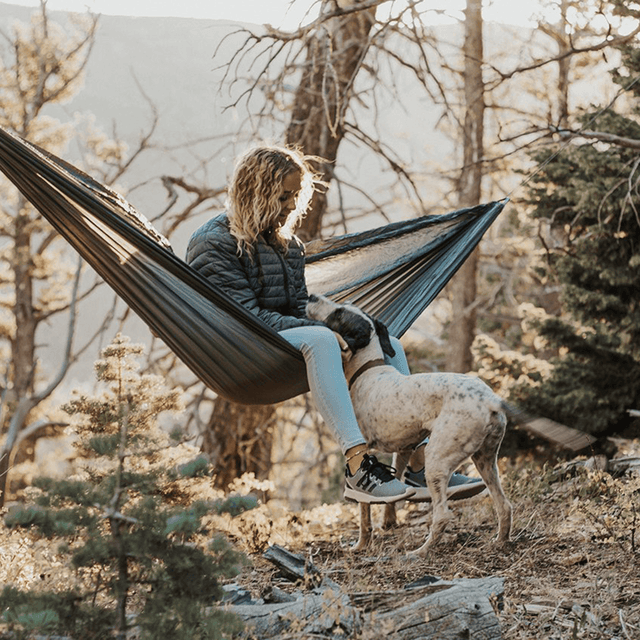 A person sitting in an Outdoor Vitals Ultralight Complete Hammock System while petting a dog outdoors in a forest.