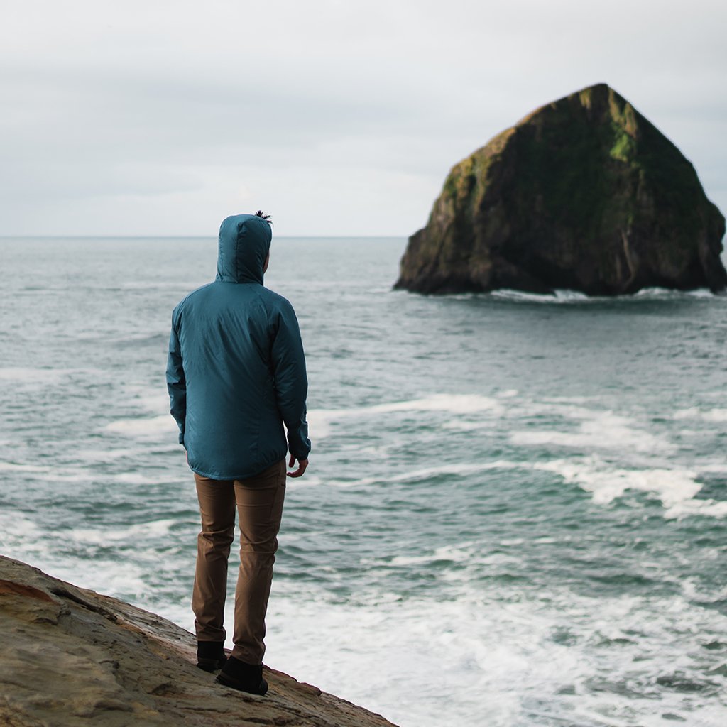 Man from behind standing on a rocky cliff overlooking the ocean and a large sea stack, wearing a Ventus Active Hoodie.