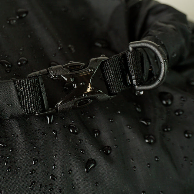 Extreme close-up of a water-resistant roll-top dry sack buckle closure with an attached D-ring, featuring water droplets on the durable coated fabric surface.