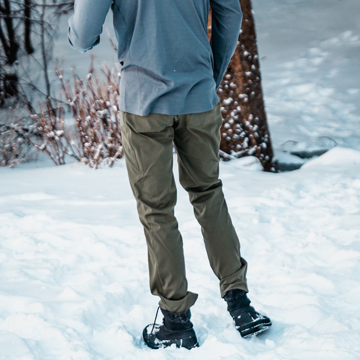Back view of a person standing in deep snow wearing Outdoor Vitals Satu Adventure Pants, winter boots, and a long-sleeve shirt outdoors.