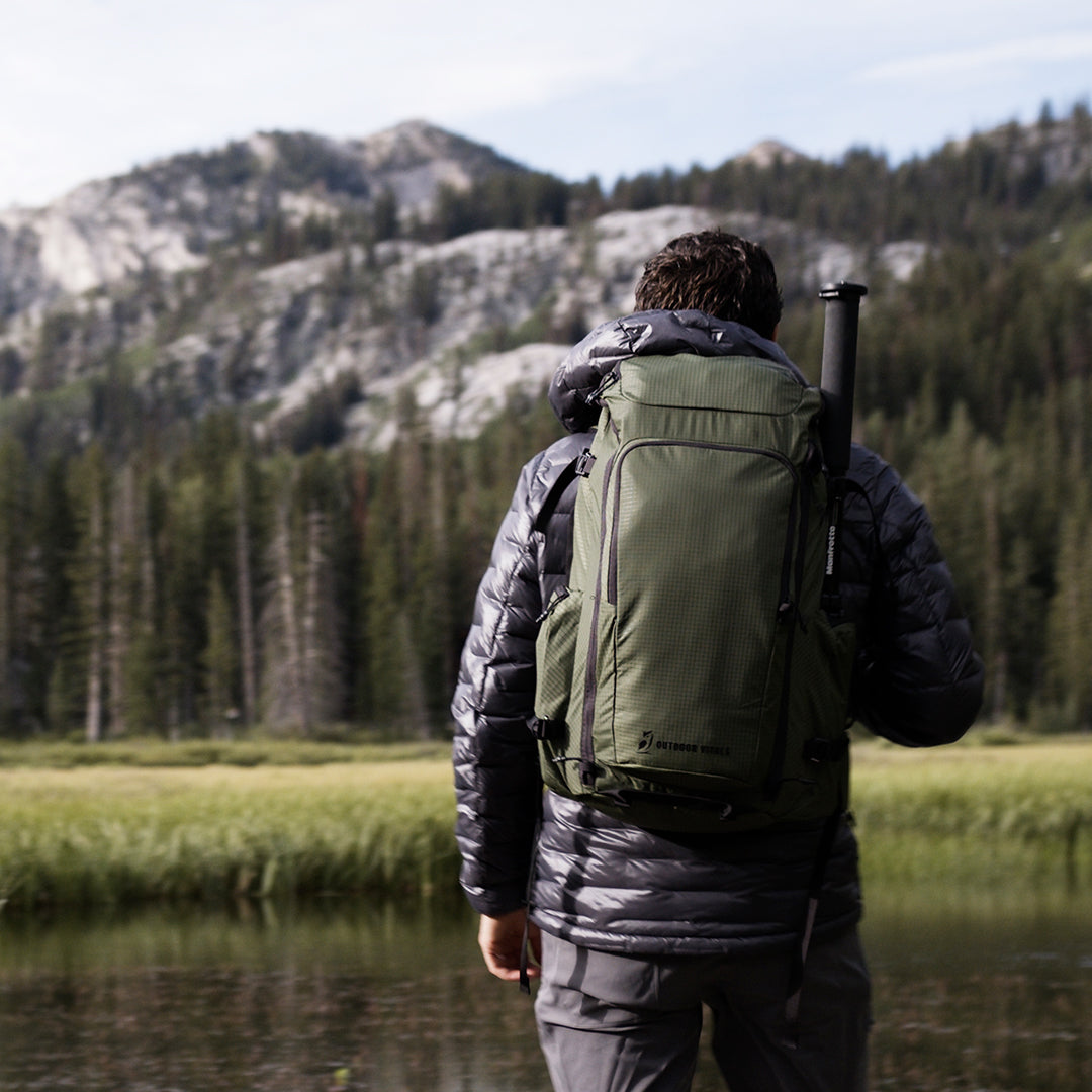 Back view of a hiker wearing a puffy jacket and the Outdoor Vitals KotaUL Ultralight Travel & Adventure Backpack with a trekking pole attached, standing near a mountain lake with pine trees and rocky peaks in the background.