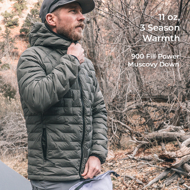 A man wearing a gray puffer jacket and a black cap stands outdoors in a wooded setting. Text on the right reads '11 oz, 3 Season Warmth' and '900 Fill Power Muscovy Down'.