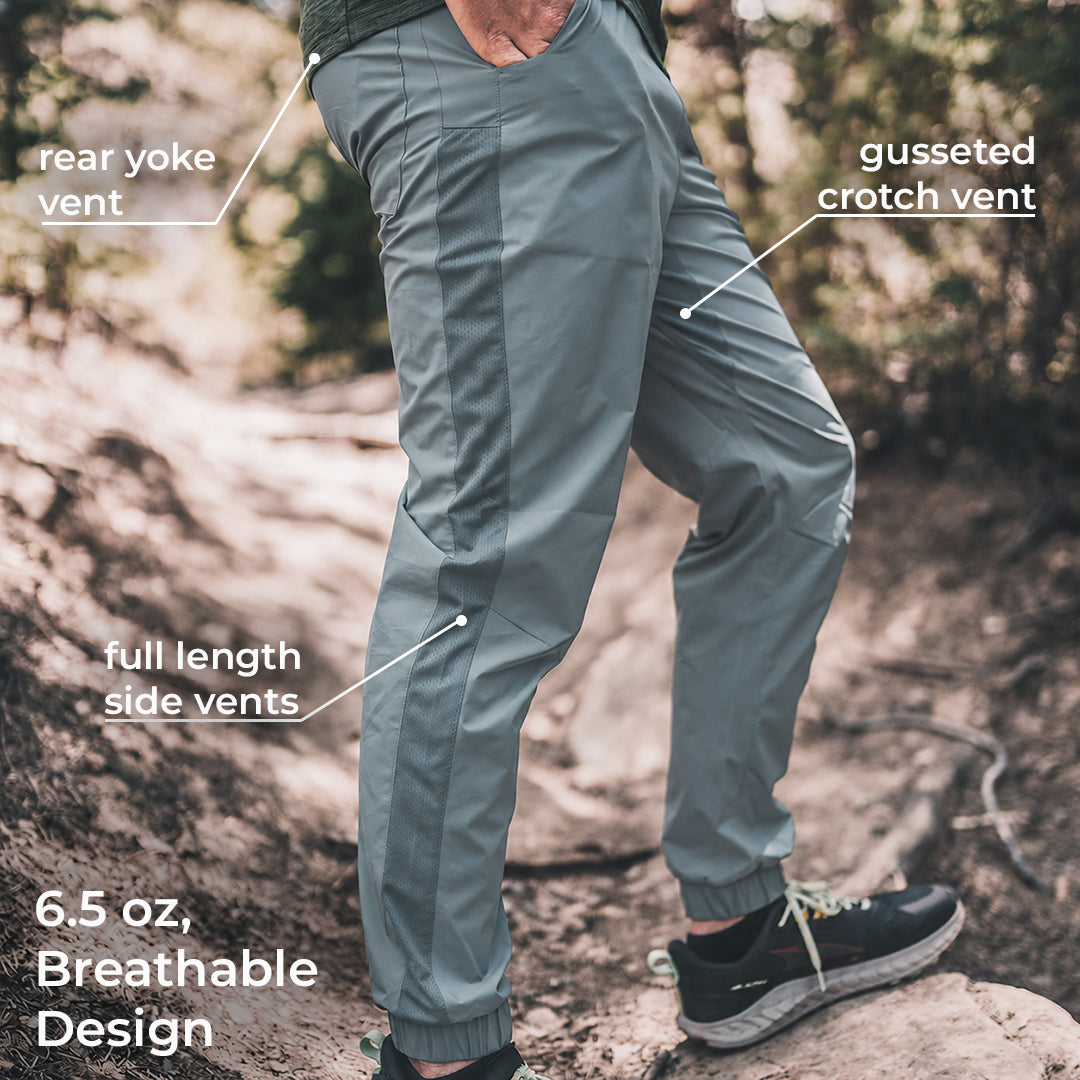 A person outdoors wearing blue technical pants with callout labels pointing to rear yoke vent, gusseted crotch vent, full length side vents, and 6.5 oz breathable design.