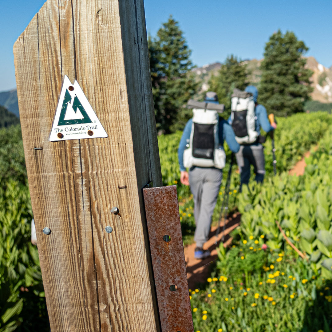 Wooden trail marker post with a Colorado Trail sign in the foreground, with two hikers wearing Outdoor Vitals CS40 Ultra Backpacks walking down a scenic mountain trail in the background.