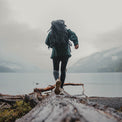 Rear view of a person wearing the Outdoor Vitals KotaUL Ultralight Travel & Adventure Backpack while balancing on a large log along a foggy lake shore with mountains in the background.