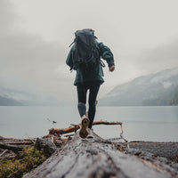 Rear view of a person wearing the Outdoor Vitals KotaUL Ultralight Travel & Adventure Backpack while balancing on a large log along a foggy lake shore with mountains in the background.