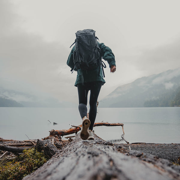 Rear view of a person wearing the Outdoor Vitals KotaUL Ultralight Travel & Adventure Backpack while balancing on a large log along a foggy lake shore with mountains in the background.