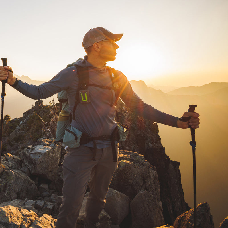 Hiker wearing the Altitude Sun Hoodie standing on a rocky peak at sunset, holding trekking poles.