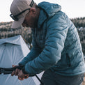 A man wearing a blue puffer jacket and a beige cap is crouching outdoors near a tent, holding tent poles in a mountain setting.