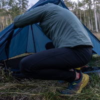 A hiker crouching outside a tent while wearing Outdoor Vitals Highline Thermal Leggings and setting up camp in a wooded area.
