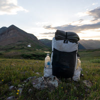 Outdoor Vitals CS40 Ultra Backpack fully packed with water bottles in the side pockets and gear strapped to the top, resting on a grassy alpine ridge with mountains in the background.