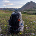 Back view of the Outdoor Vitals CS40 Ultra Backpack standing in an alpine meadow, displaying shoulder straps, hip belt pockets, attached satellite messenger device, and side water bottle pocket with scenic mountain peaks in the background.