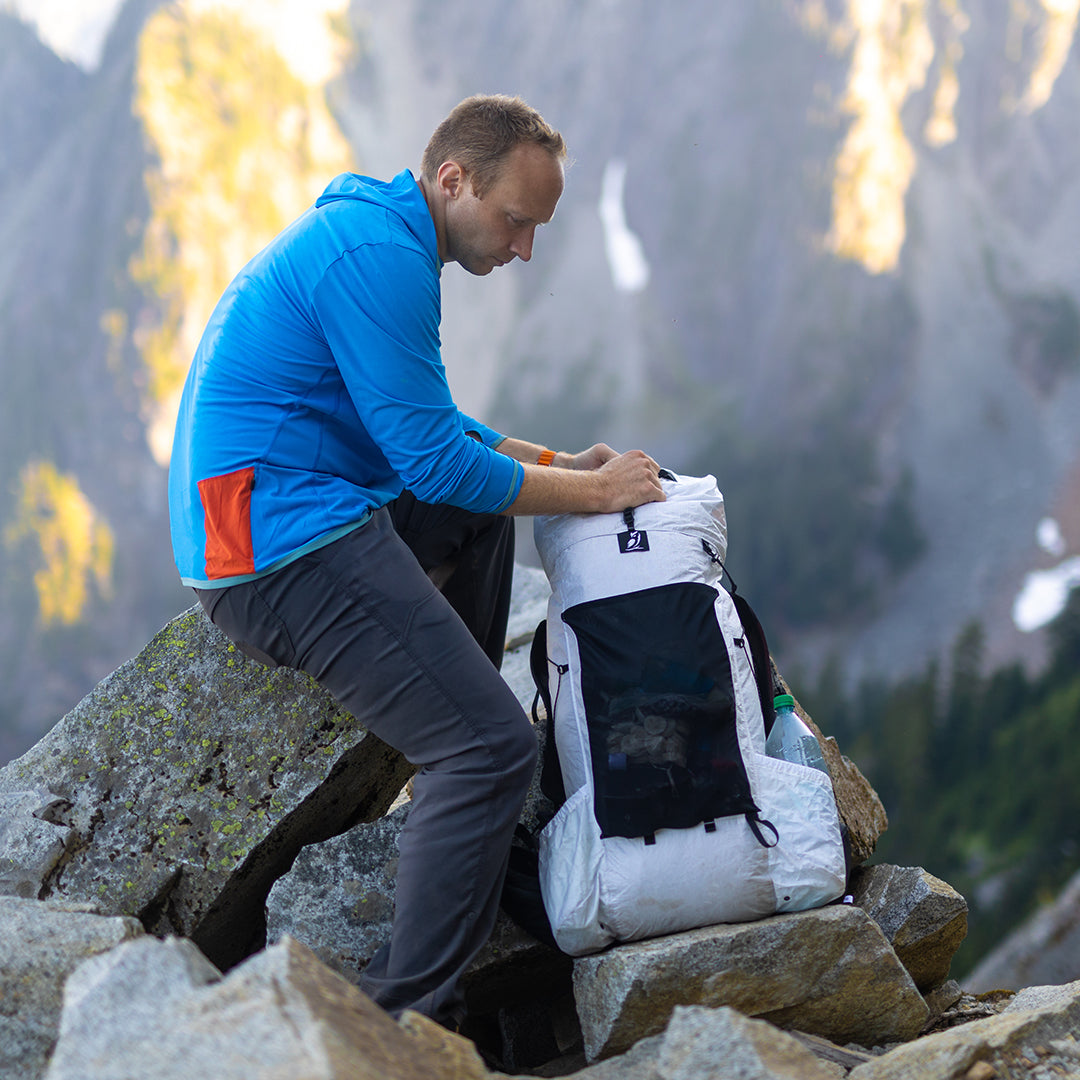 Side view of a hiker sitting on rocks and adjusting the roll-top closure of the Outdoor Vitals CS40 Ultra Backpack on a mountain ridge.