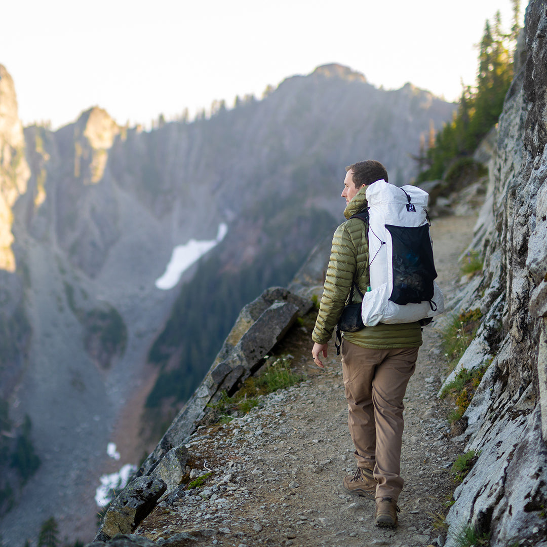 Back view of a hiker carrying the Outdoor Vitals CS40 Ultra Backpack while trekking along a narrow mountain cliff path overlooking a deep valley.