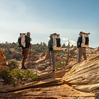 Three hikers standing on a rocky desert overlook, all wearing Outdoor Vitals CS40 Ultra Backpacks while using trekking poles during a backcountry trek.