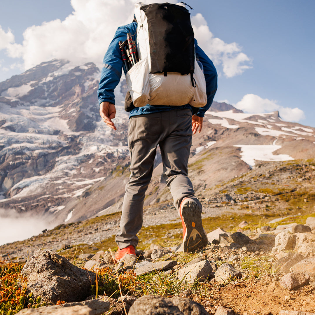 Back view of a hiker walking up a rocky alpine trail carrying the Outdoor Vitals CS40 Ultra Backpack with a tripod stowed in the side pocket and snow-covered peaks in the background.