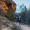 Rear view of two backpackers wearing Outdoor Vitals CS40 Ultra Backpacks as they hike along a sandy trail below a steep rock cliff face in a canyon environment.