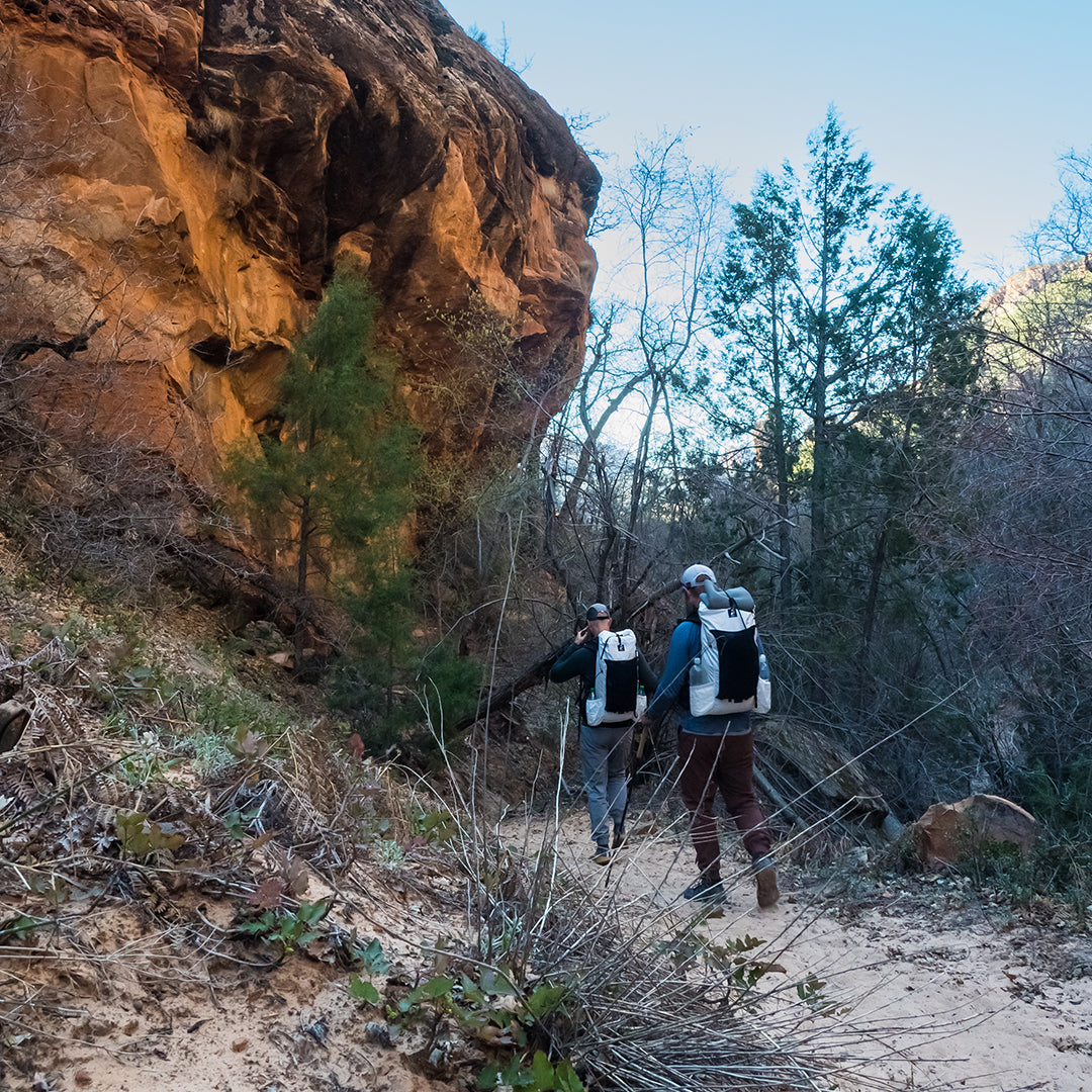 Rear view of two backpackers wearing Outdoor Vitals CS40 Ultra Backpacks as they hike along a sandy trail below a steep rock cliff face in a canyon environment.
