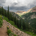 Distant view of a backpacker carrying the Outdoor Vitals CS40 Ultra Backpack while hiking along a rocky mountain ridge trail with pine trees and peak landscape views.