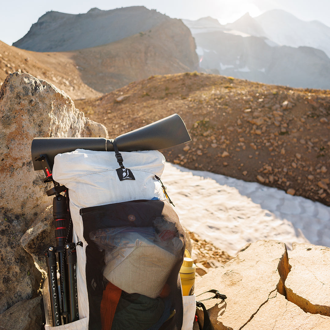 Close-up view of the Outdoor Vitals CS40 Ultra Backpack resting against a rock in a high-alpine landscape, fully loaded with a sleeping pad strapped on top, tripod attached to the side, and gear packed in the front mesh pocket.