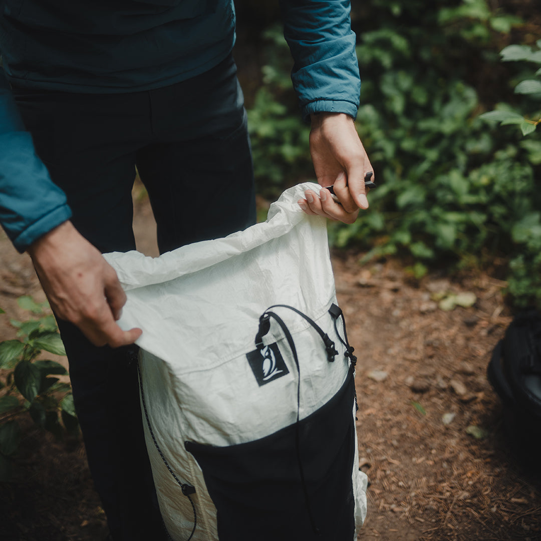 Close-up of a person unrolling the top closure of the Outdoor Vitals CS40 Ultra Backpack while standing on a forest trail.