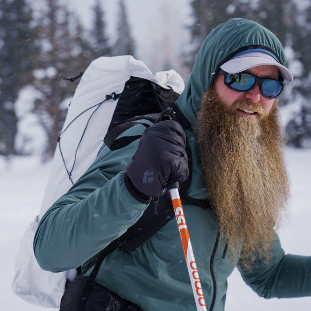 Close-up profile of a bearded winter hiker wearing a hooded jacket, gloves, and sunglasses while trekking through snow with poles and the Outdoor Vitals CS40 Ultra Backpack.