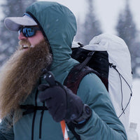 Side profile of a bearded winter hiker smiling while wearing a hooded jacket, cap, and the Outdoor Vitals CS40 Ultra Backpack during a snowstorm on a mountain trail.