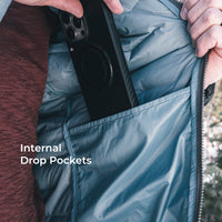 Close-up of a person opening a blue-gray puffer jacket to show an internal drop pocket holding a black smartphone. White text on the image reads 'Internal Drop Pockets'.