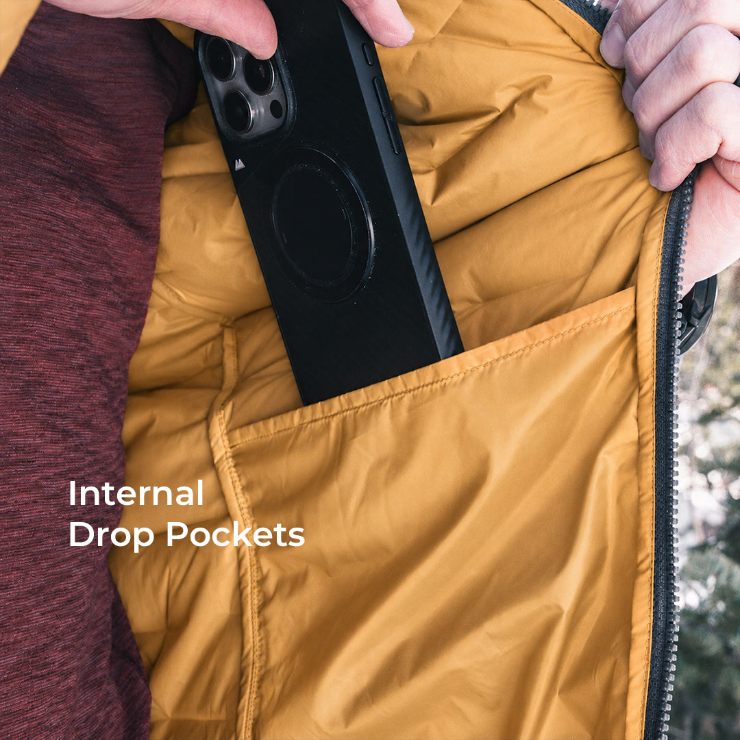Close-up of a person's hands opening a mustard yellow puffer jacket to reveal an internal drop pocket holding a black smartphone. White text on the image reads 'Internal Drop Pockets'.