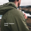 A man viewed from behind wearing an olive green hooded jacket covered in water droplets, with a blurred outdoor coastal landscape in the background. White text on the lower left reads 'DWR Treated Shell'.