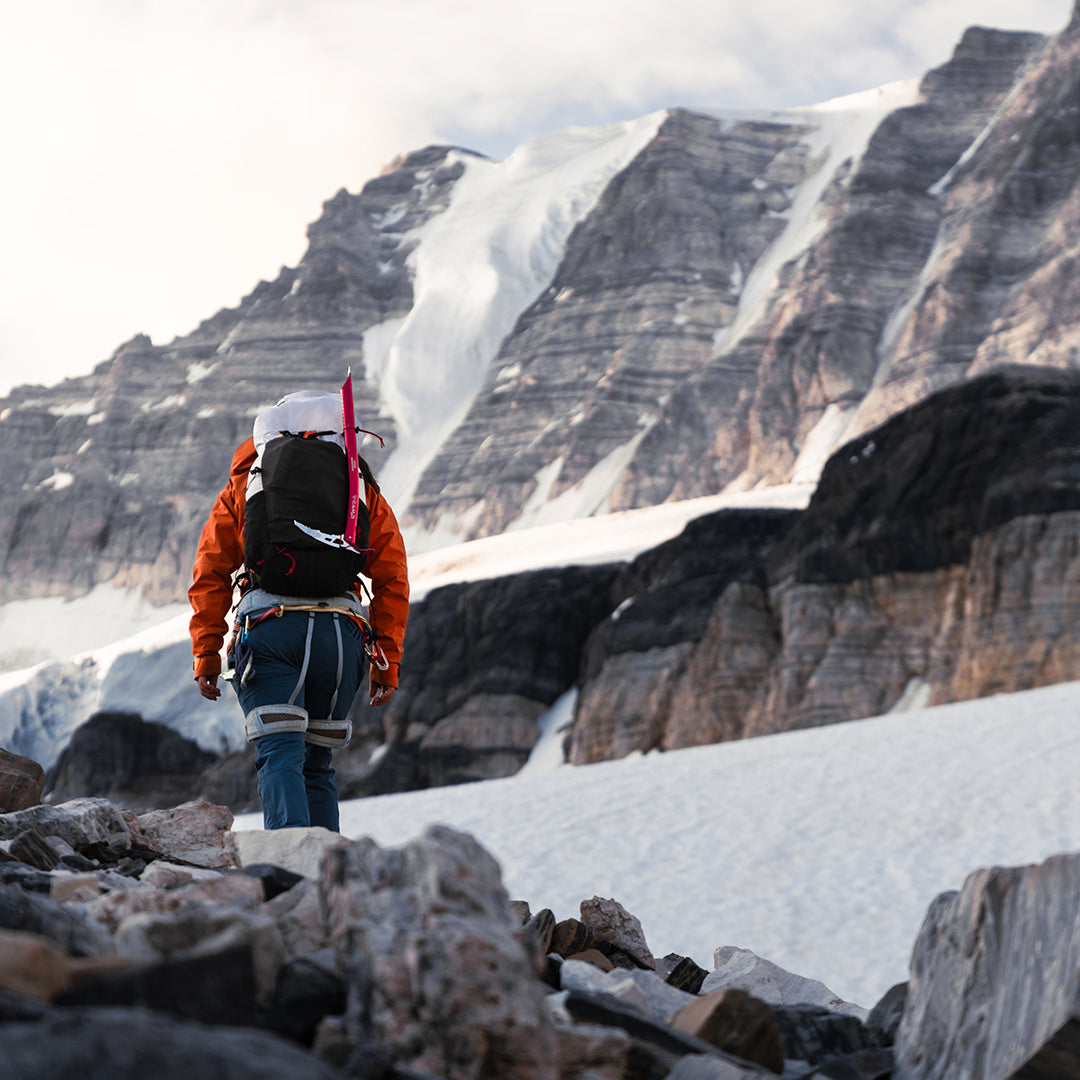 Rear view of a mountaineer trekking toward a snow-covered alpine peak while wearing the Outdoor Vitals Skyline 30 Fastpack, equipped with an ice axe strapped to the side attachment loops.