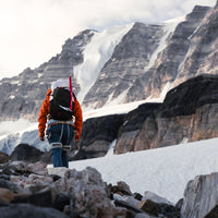 Rear view of a mountaineer trekking toward a snow-covered alpine peak while wearing the Outdoor Vitals Skyline 30 Fastpack, equipped with an ice axe strapped to the side attachment loops.
