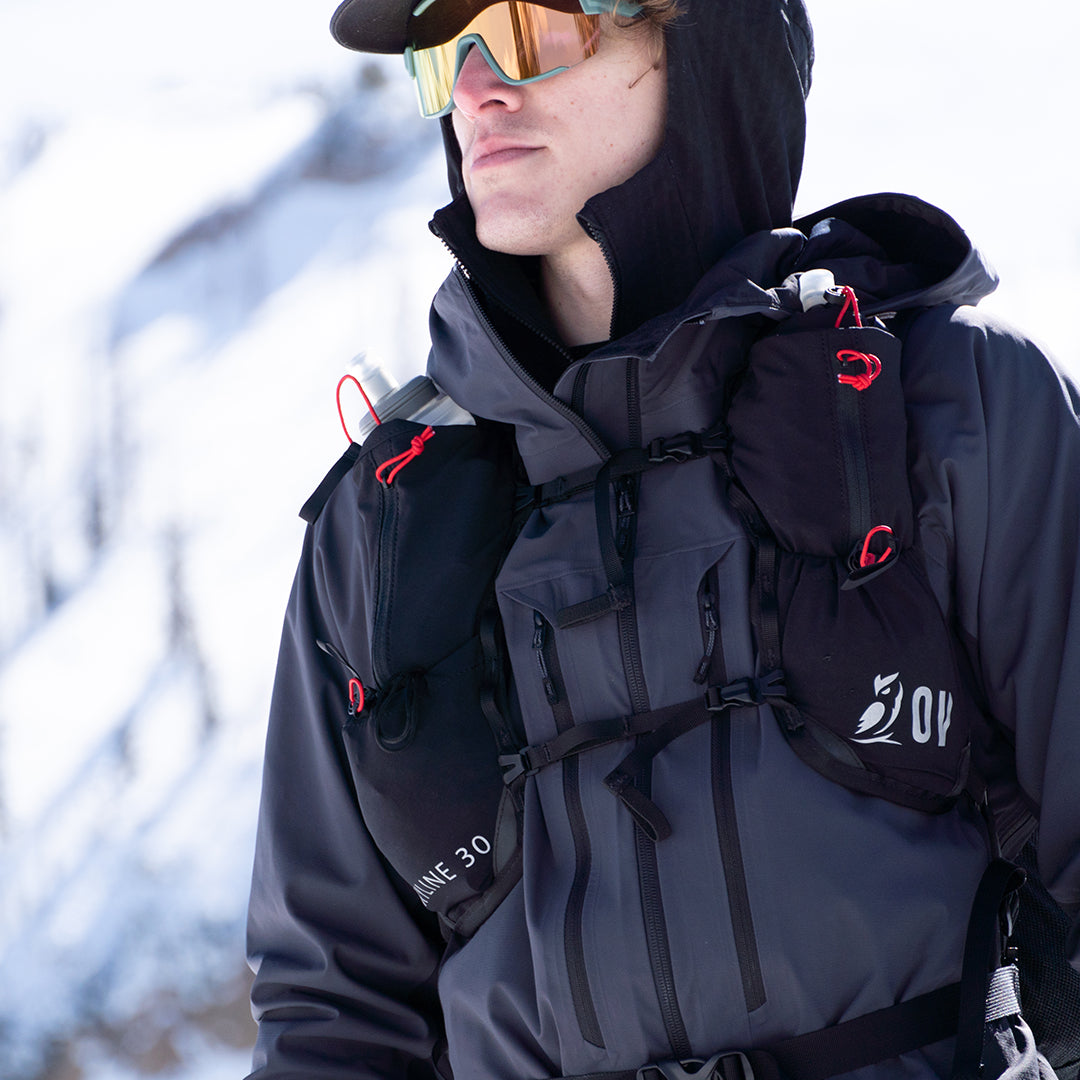Close-up front view of a mountaineer in snowy conditions wearing the Outdoor Vitals Skyline 30 Fastpack vest harness, featuring integrated flask pockets, zippered mesh compartments, and dual sternum straps.